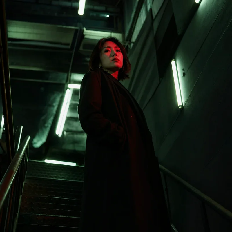 Retrato de una mujer con abrigo negro en una escalera, iluminada con luz verde y roja